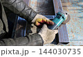 A close-up of a workers hands in thick protective gloves. Using a turquoise-colored handheld angle grinder, the craftsman carefully processes a welded joint between rectangular metal pipes. 144030735