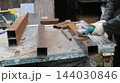 A worker wearing gloves uses an angle grinder to clean the surface of a rectangular metal profile. On an old workbench, workpieces showing signs of corrosion are laid out, ready for assembly. 144030846