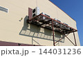 A metal framework holding the outdoor units of air conditioning systems is mounted on the beige facade of a commercial building. On a sunny day, the latticed structure casts a sharp shadow 144031236
