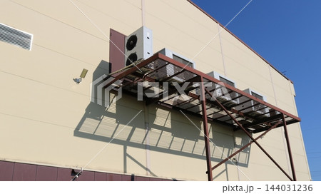 A metal framework holding the outdoor units of air conditioning systems is mounted on the beige facade of a commercial building. On a sunny day, the latticed structure casts a sharp shadow 144031236