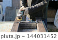 A craftsman wearing a leather jacket and protective gloves works on metal using an angle grinder. He carefully grinds the welded joints of rectangular profile pipes secured to a workbench. 144031452