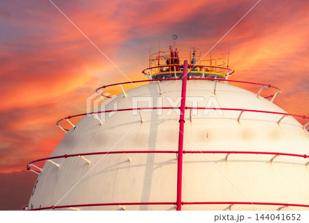 Spherical gas storage tank with red piping beneath an orange cloudy sky at a petrochemical facility, illustrating industrial fuel storage and energy infrastructure with a dramatic atmospheric backdrop Spherical gas storage tank with red piping beneath an orange cloudy sky at a petrochemical facility, illustrating industrial fuel storage and energy infrastructure with a dramatic atmospheric backdrop 144041652