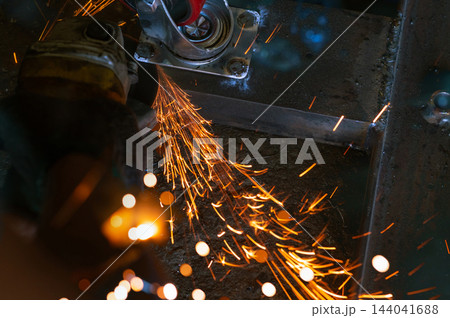 Worker grinding a steel frame with an angle grinder beside a caster wheel mounting plate, orange sparks streaming across the work surface during metal fabrication and industrial component finishing Worker grinding a steel frame with an angle grinder beside a caster wheel mounting plate, orange sparks streaming across the work surface during metal fabrication and industrial component finishing 144041688