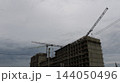 The construction process of a multi-story reinforced concrete building. Against a backdrop of gray clouds, the structural framework under construction rises upward, alongside the booms of two cranes  144050496