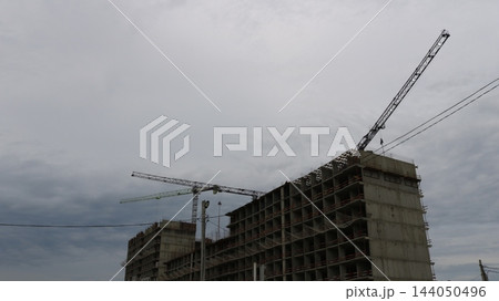 The construction process of a multi-story reinforced concrete building. Against a backdrop of gray clouds, the structural framework under construction rises upward, alongside the booms of two cranes The construction process of a multi-story reinforced concrete building. Against a backdrop of gray clouds, the structural framework under construction rises upward, alongside the booms of two cranes 144050496