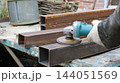 A gloved hand holds an angle grinder, cleaning the surface of a rectangular metal profile. Small sparks fly from the rotating disc, while other stacked workpieces are visible in the background. 144051569