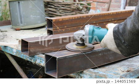 A gloved hand holds an angle grinder, cleaning the surface of a rectangular metal profile. Small sparks fly from the rotating disc, while other stacked workpieces are visible in the background. 144051569