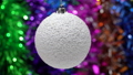 Spinning white Xmas ball on blurry bokeh background of colored and shining tinsel, holiday lights. Closeup view of hanging single holiday Christmas ball. Motion blur. Selective focus on foreground. 84764333