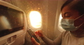 Woman using phone on plane. Young woman holding smartphone sitting in airplane. Sunset and flare in window seat. Hands swiping on screen. Asian Caucasian multiracial model wearing face mask 86769032