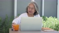 Happy elderly Asian woman with glasses using laptop at her desk in backyard and drinking Orange juice, smiling old mature lady paying bill with online application on laptop 94552534