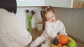 Beautiful mother and daughter spend time together in a white cozy kitchen. Mother feeds her daughter tangerine. Mother's Day 100658692